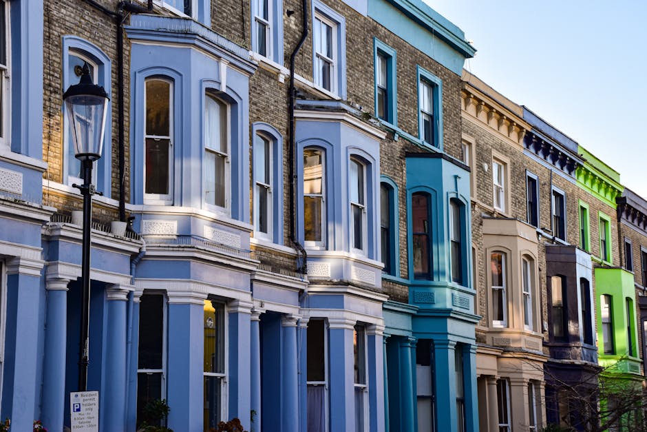 A row of multi-storey terraced houses with elaborate Victorian-style facade details, including bay windows and decorative mouldings, situated along a street in Westminster SW1A. The buildings are painted in various pastel and neutral shades, such as light blue, beige, and green, with some painted accents highlighting architectural features. A black lamppost with a traditional lantern stands on the pavement in the foreground. The street scene is captured during daylight, with clear sky reflected in the windows. The buildings appear well-maintained, and the scene depicts a typical urban residential area suitable for house removals and furniture transport. The presence of open windows and an orderly environment suggests the possibility of a home relocation or packing and moving activities, with [COMPANY_NAME] providing removals services in Westminster.