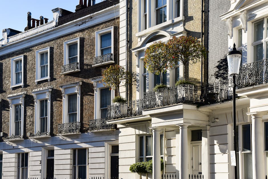 A multi-storey residential building with a white facade and black iron balcony railings, located on a street in Westminster. Several balconies feature potted trees and plants. The ground floor has a white entrance door beside a classic black lamppost. Sunlight casts shadows on the building's exterior, highlighting the window frames and architectural details. The scene depicts the exterior of an urban property, suitable for home relocation or furniture transport, with a focus on the building's facade and exterior environment, supporting house removals and packing processes. As part of a professional moving service, the image suggests the early stages of a relocation, such as loading or preparatory activities at the property, with a clear view of the street and building frontage.