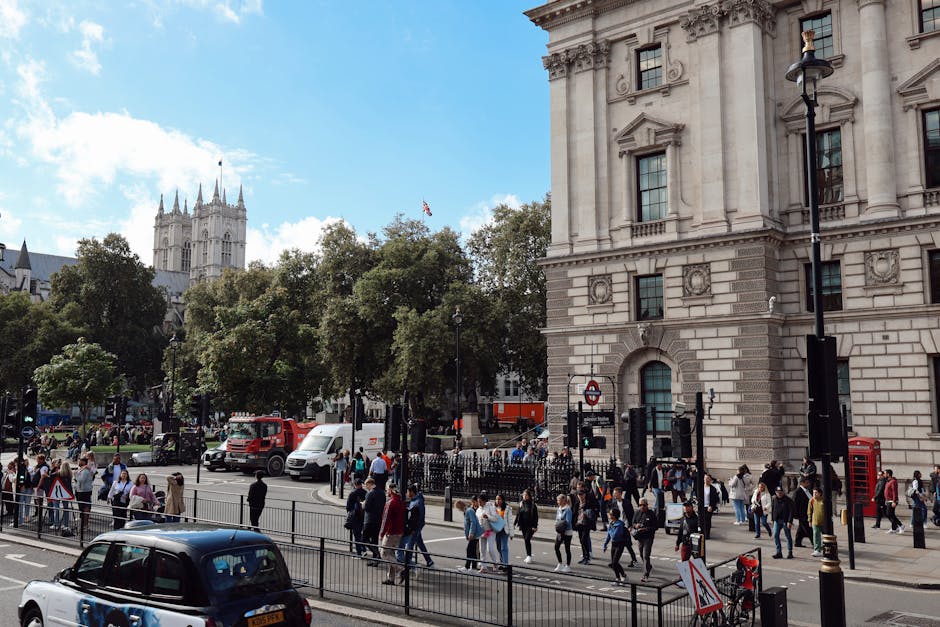 A street scene in Westminster featuring the Palace of Westminster and the Elizabeth Tower, known as Big Ben, under a partly cloudy sky. In the foreground, a man with a furniture trolley is loading or