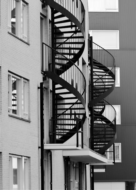 A black and white photograph showing a fire escape spiral staircase installed on the exterior of a modern residential building with multiple story levels. The staircase is constructed of metal with ra